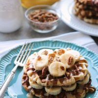 Cinnamon Roll Waffles are topped with freshly sliced bananas, crunchy nuts, and a maple cream cheese drizzle.