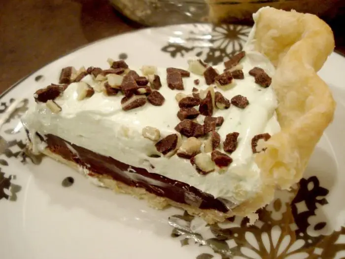 Slice of chocolate mint pie on a plate with layers of pastry crust, chocolate ganache, mint no bake cheesecake, and chopped Andes mints.