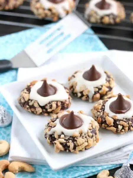 Chocolate cookie dough is rolled in nuts and topped with buttercream and chocolate kisses to create these thick and chewy Chocolate Cream Thumbprint Cookies.