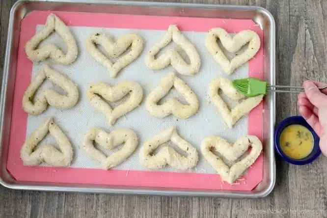 Brushing egg wash onto heart shaped breadsticks before baking.