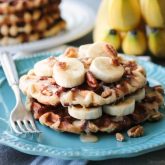 Cinnamon Roll Waffles are topped with freshly sliced bananas, crunchy nuts, and a maple cream cheese drizzle.