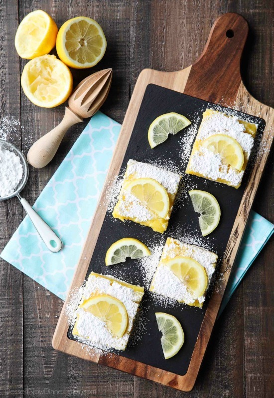 Slices of lemon bars on a board with powdered sugar and lemon slices on top.