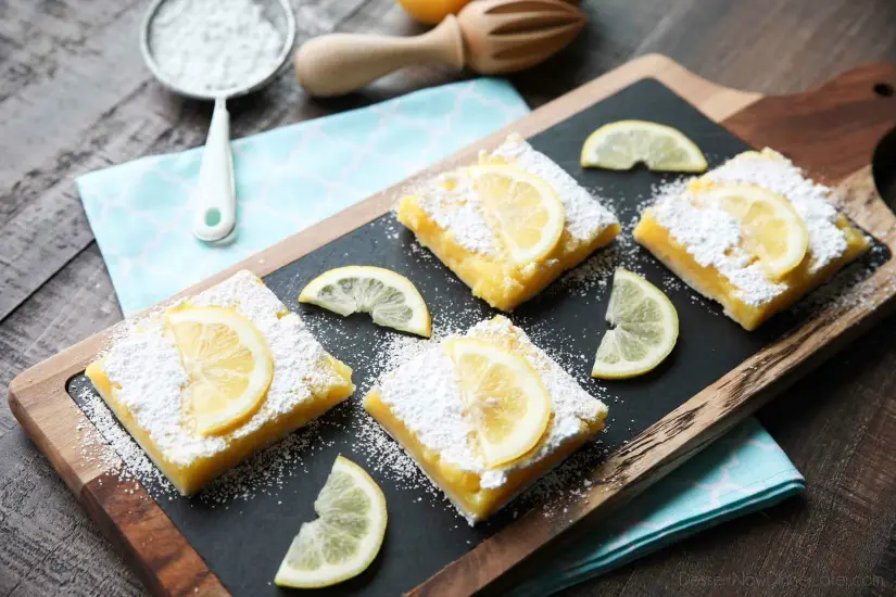 Slices of lemon bars on a board with powdered sugar and lemon slices on top.