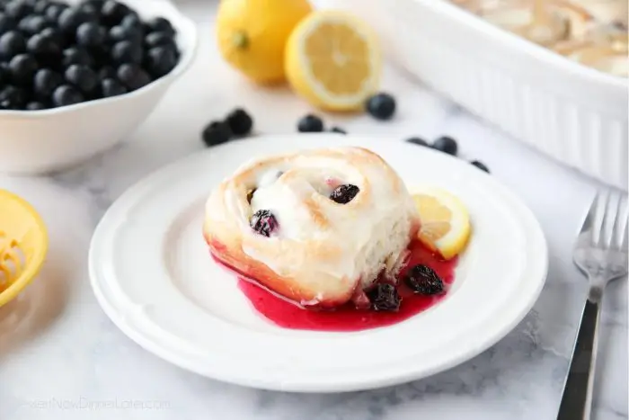 A lemon blueberry sweet roll on a plate with blueberry sauce and a slice of lemon.