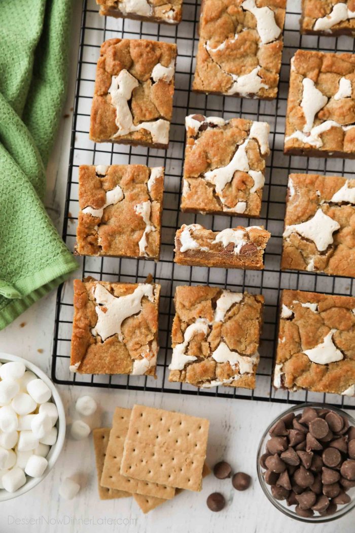 Gooey s'mores bars on a cooling rack with ingredients of graham crackers, chocolate chips, and mini marshmallows nearby. | Dessert Now Dinner Later