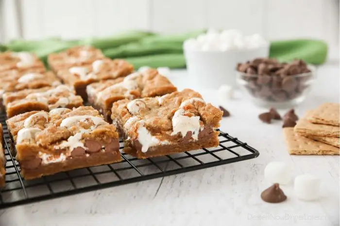 Gooey s'mores bars on a cooling rack with ingredients of graham crackers, chocolate chips, and mini marshmallows nearby. | Dessert Now Dinner Later