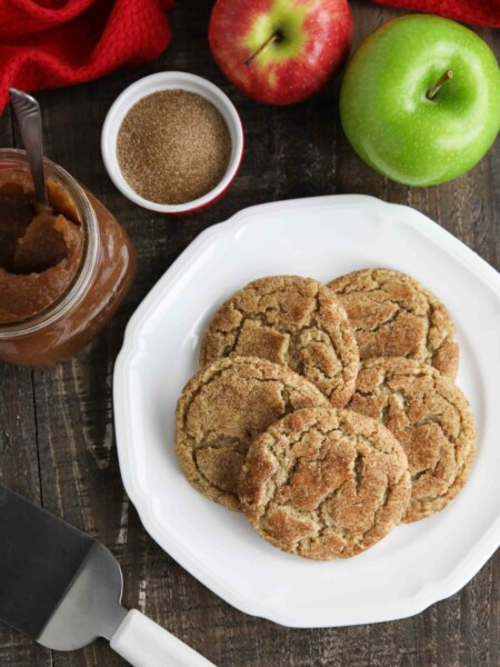 Crinkle topped apple butter cookies on plate with apple butter, cinnamon sugar, and fresh apples nearby.