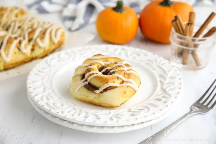 A Pumpkin Pie Cinnamon Roll on a plate with decorative pumpkins and cinnamon sticks in the background.