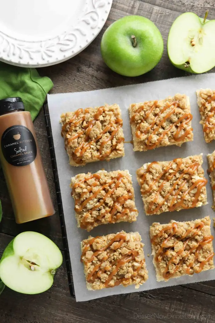 Squares of caramel apple cheesecake bars on parchment paper surrounded by Granny Smith apples and a bottle of salted caramel.