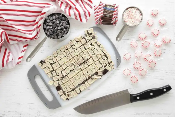 Peppermint bark squares chopped on a cutting board with a chef's knife on the side. Measuring cups full of chocolate chips and crushed peppermint candies.