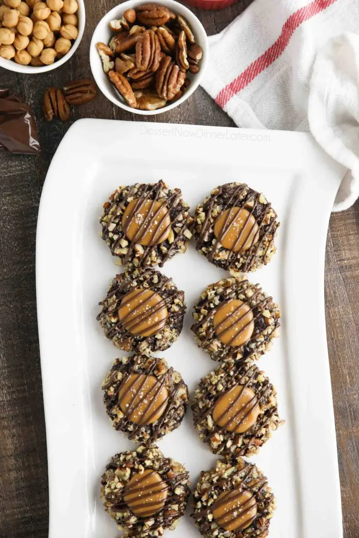 Long white plate of turtle thumbprint cookies with pecans and caramel bits nearby.