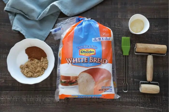 A package of Rhodes frozen White Bread loaves with a bowl of sugar and cinnamon, a pastry brush, mini rolling pin, and petite bowl of melted butter on the sides.