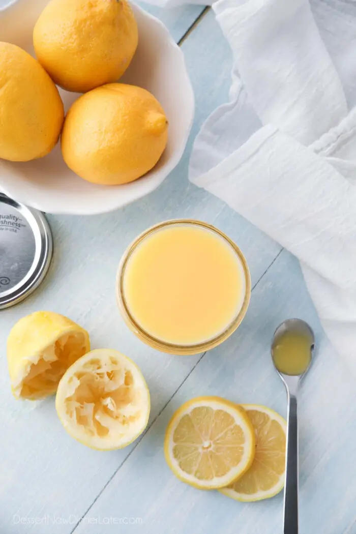 Lemon curd in a jar surrounded by whole lemons, juiced lemons, and lemon slices.