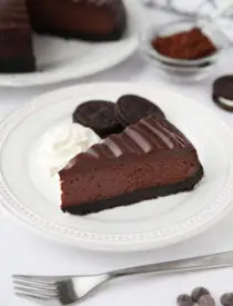 A slice of Triple Chocolate Cheesecake on a plate with Oreos and whipped cream on the side.