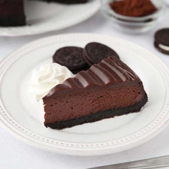 A slice of Triple Chocolate Cheesecake on a plate with Oreos and whipped cream on the side.