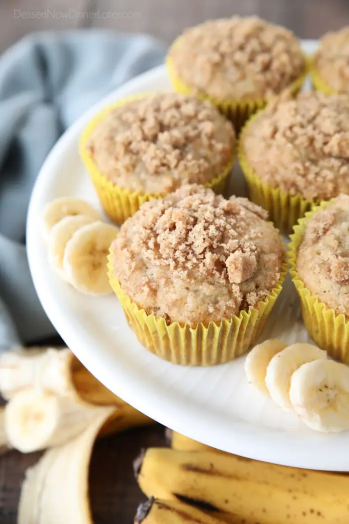 White cake plate with banana muffins in yellow wrappers with crumb streusel on top and slices of banana on the sides.