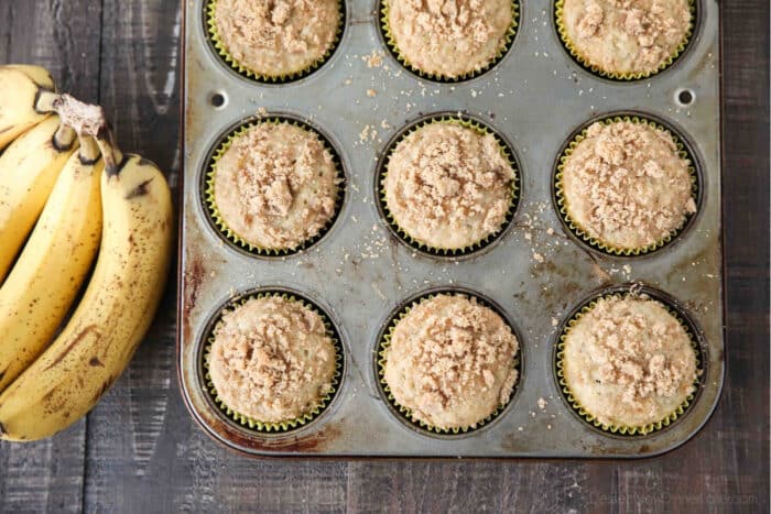 Baked banana crumb muffins in cupcake tin with bunch of bananas on the side.