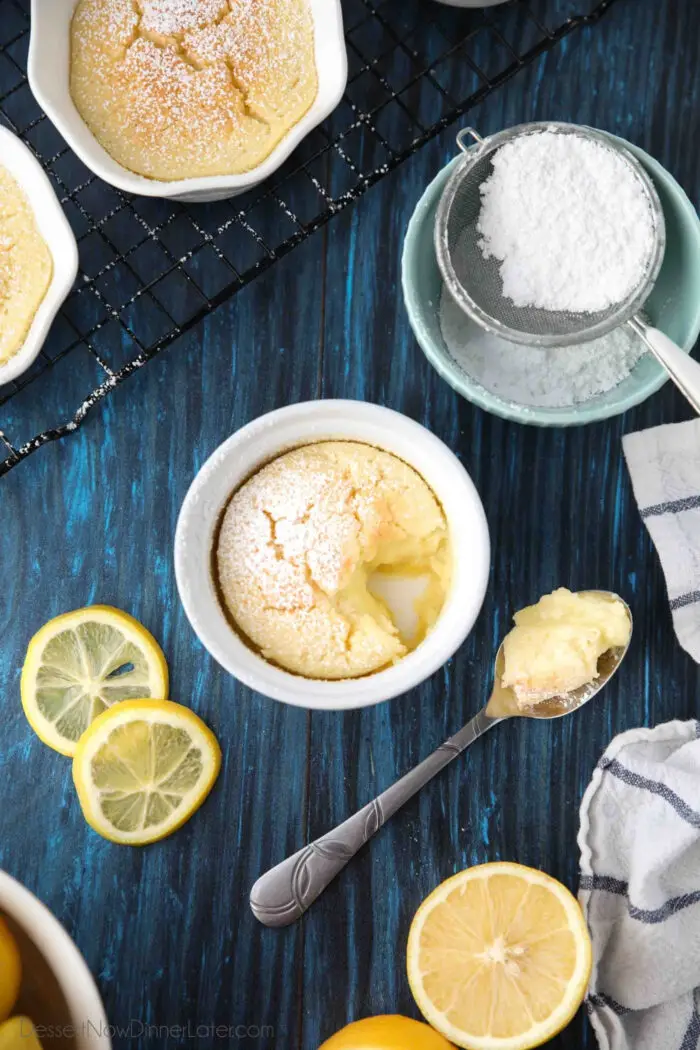 Spoonful of lemon pudding cake next to ramekin with lemons and powdered sugar on the side.