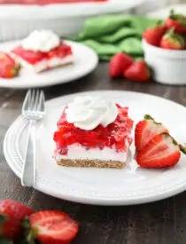 A square of strawberry delight on a plate. (A layered dessert with a graham cracker crust, no bake cheesecake filling, and a layer of fresh strawberries and jello with a swirl of whipped cream on top.)
