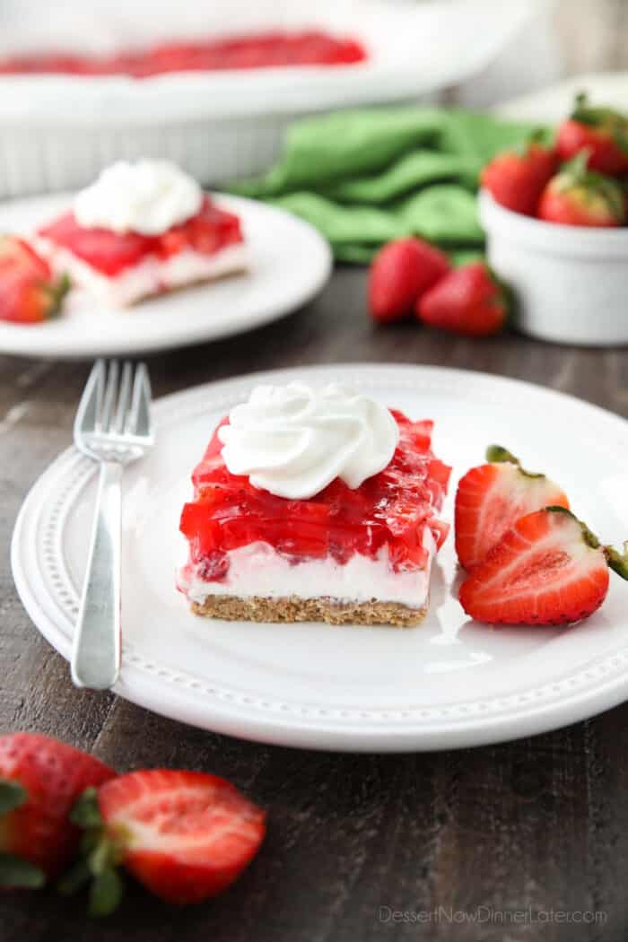 A square of strawberry delight on a plate. (A layered dessert with a graham cracker crust, no bake cheesecake filling, and a layer of fresh strawberries and jello with a swirl of whipped cream on top.)
