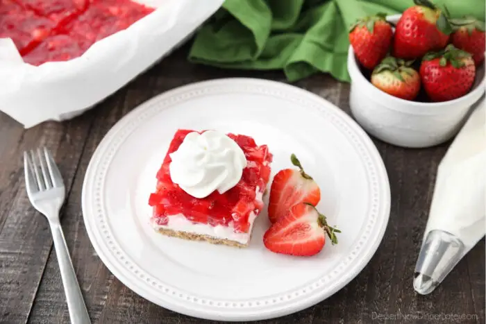 A piece of strawberry delight on a plate with a halved strawberry on the side.