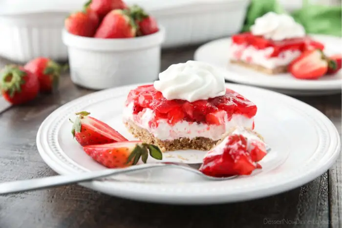 A square of strawberry delight on a plate. A layered dessert with a graham cracker crust, no bake cheesecake filling, and a layer of fresh strawberries and jello with a swirl of whipped cream on top.