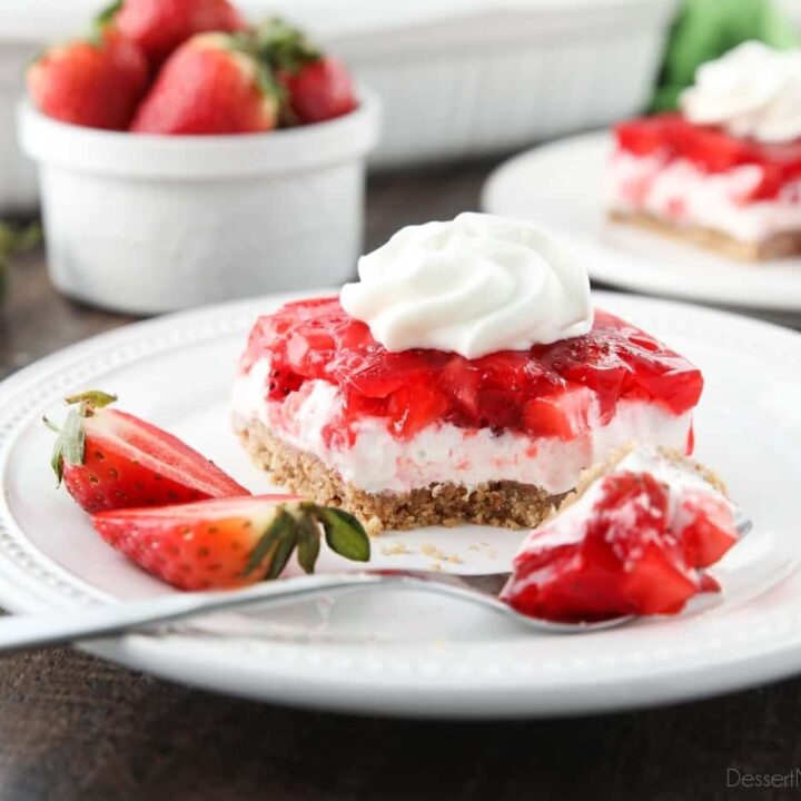 A square of strawberry delight on a plate. A layered dessert with a graham cracker crust, no bake cheesecake filling, and a layer of fresh strawberries and jello with a swirl of whipped cream on top.