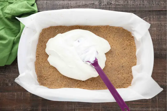 Cooked and cooled graham cracker crust in a 13x9-inch pan with no bake cheesecake being spread over it with a spatula.