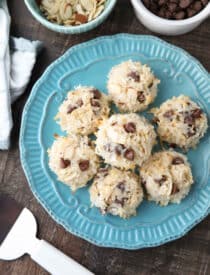 Top view of Almond Joy Cookies (chocolate almond macaroons) on a plate with bowls of chocolate chips and sliced almonds on the side.