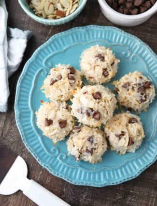 Top view of Almond Joy Cookies (chocolate almond macaroons) on a plate with bowls of chocolate chips and sliced almonds on the side.