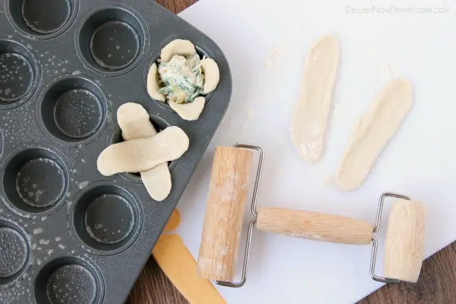 A mini rolling pin next to two logs of dough rolled flat. A mini muffin pan with two pieces of flattened criss-crossed dough in one cup. Artichoke dip inside another cup of criss-crossed dough.
