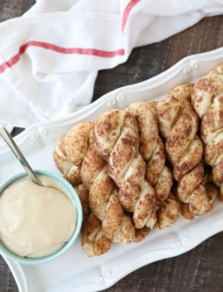 Twisted cinnamon sugar breadsticks on a plate with a bowl of cream cheese glaze.