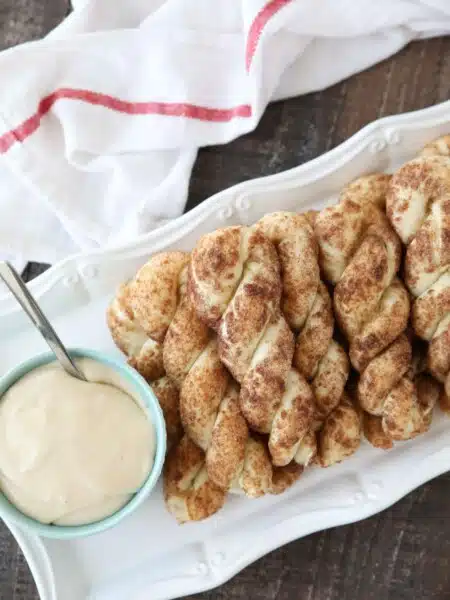 Twisted cinnamon sugar breadsticks on a plate with a bowl of cream cheese glaze.