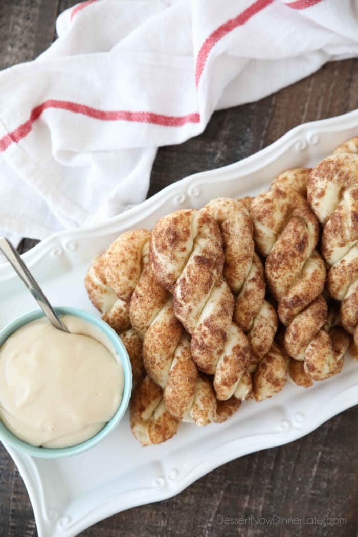 Twisted cinnamon sugar breadsticks on a plate with a bowl of cream cheese glaze.