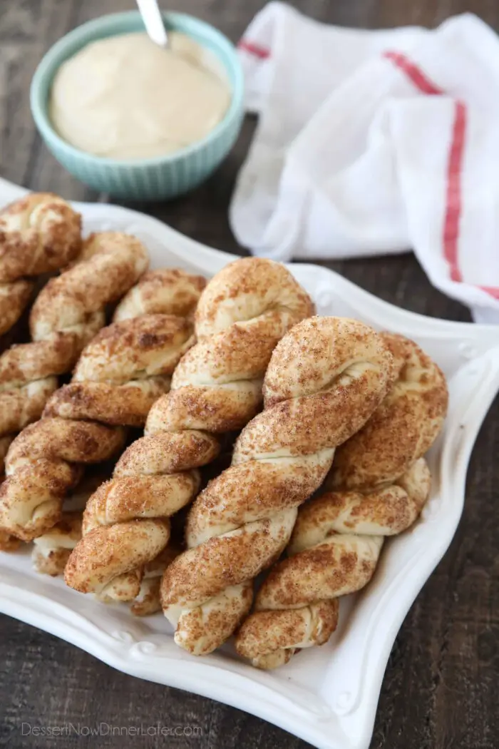 Focus on plate of cinnamon breadsticks with bowl of cream cheese dip and a towel in the background.