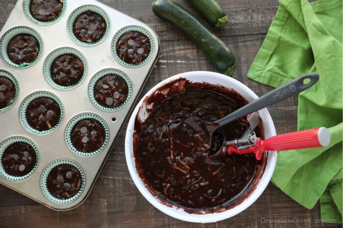 Chocolate Zucchini Muffin batter in a bowl with a spatula and scoop next to a muffin tray with paper liners and batter scooped inside.