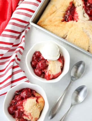 Two bowls of easy cherry cobbler with two spoons on the side. One serving of cobbler has ice cream on top, and is sitting next to the pan it was baked in.