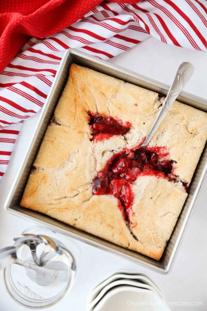 Cherry Cobbler in a square baking pan with a spoon revealing cherry pie filling underneath the cake.