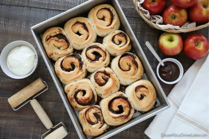 Baked Apple Butter Cinnamon Rolls in Pan with cream cheese glaze in a bowl on the side.