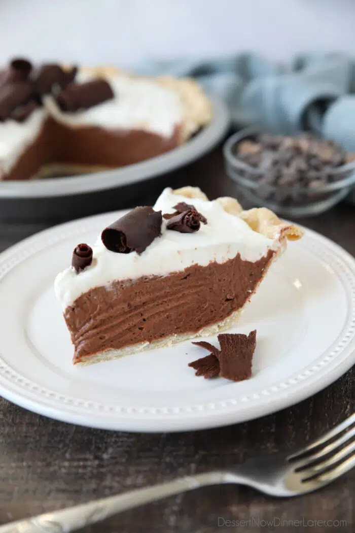 Slice of pie on a plate with a flaky pastry crust layered with chocolate pudding filling, whipped cream, and chocolate curls on top.