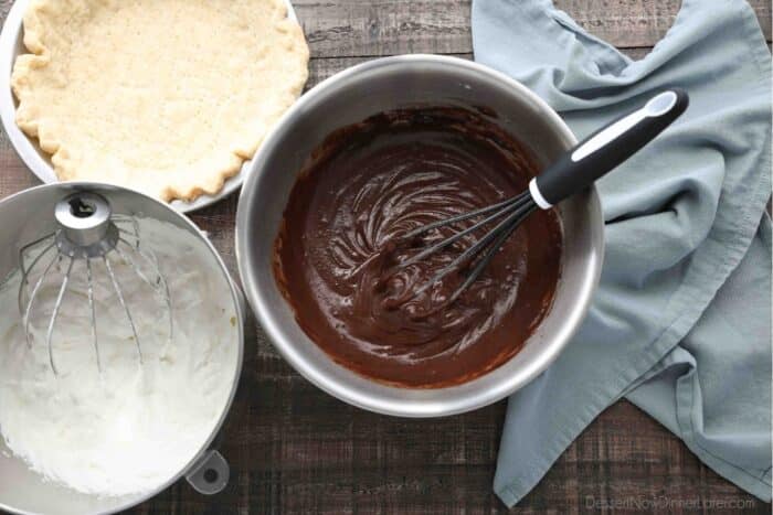 A pan with a baked pie crust, a bowl of sweetened whipped cream, and a bowl of chocolate pudding with a whisk.