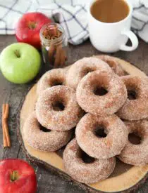 Baked Apple Cider Donuts stacked on top of a wooden cutting board with a cup of cider, fresh apples, and cinnamon sticks on the side.