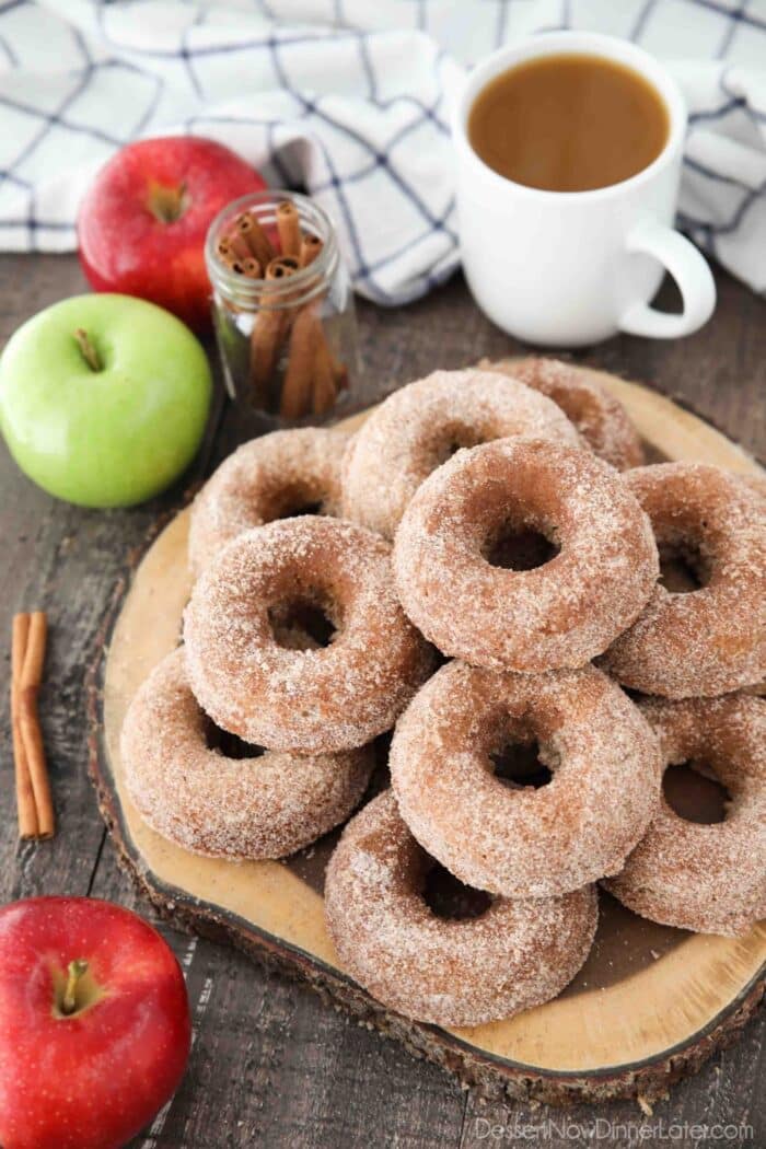 Baked Apple Cider Donuts stacked on top of a wooden cutting board with a cup of cider, fresh apples, and cinnamon sticks on the side.