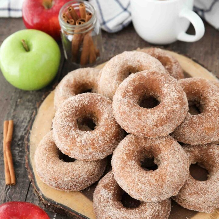 Baked Apple Cider Donuts stacked on top of a wooden cutting board with a cup of cider, fresh apples, and cinnamon sticks on the side.