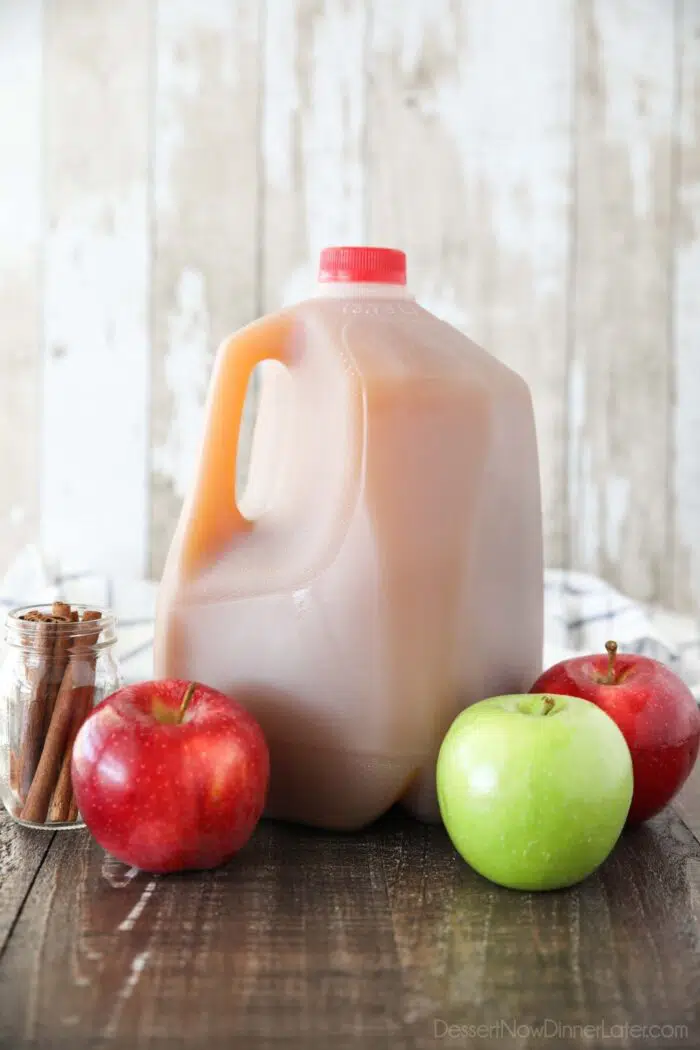 Gallon jug of fresh apple cider with apples and cinnamon sticks surrounding it.