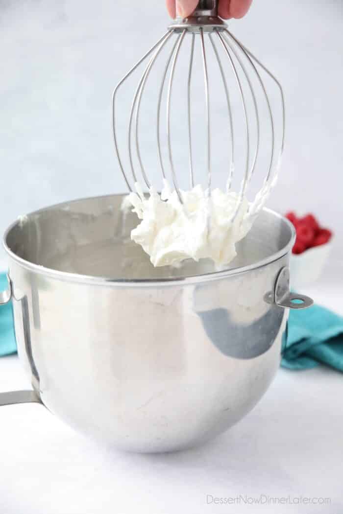 Pulling whisk attachment out of mixing bowl with whipped cream frosting on it.