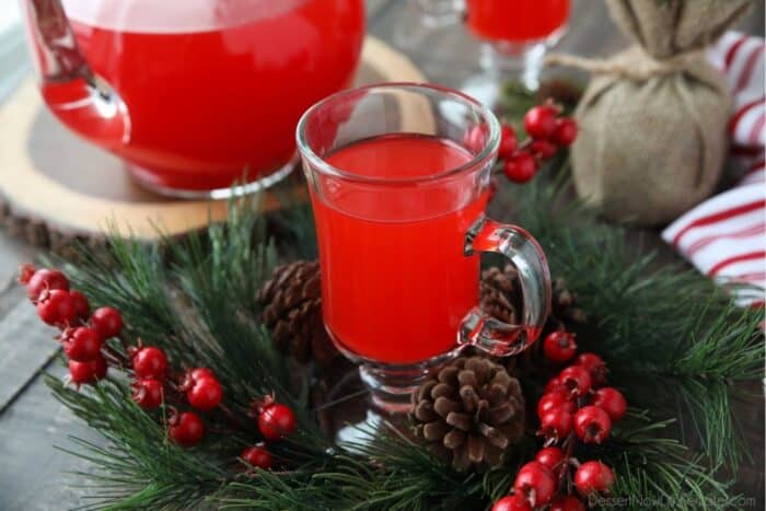 A cup of red Christmas punch with pine leaves, holly berries, and pine cones surrounding it.