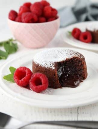 Molten chocolate lava cake on plate dusted with powdered sugar and a side of raspberries and mint, with gooey center melting onto the plate.