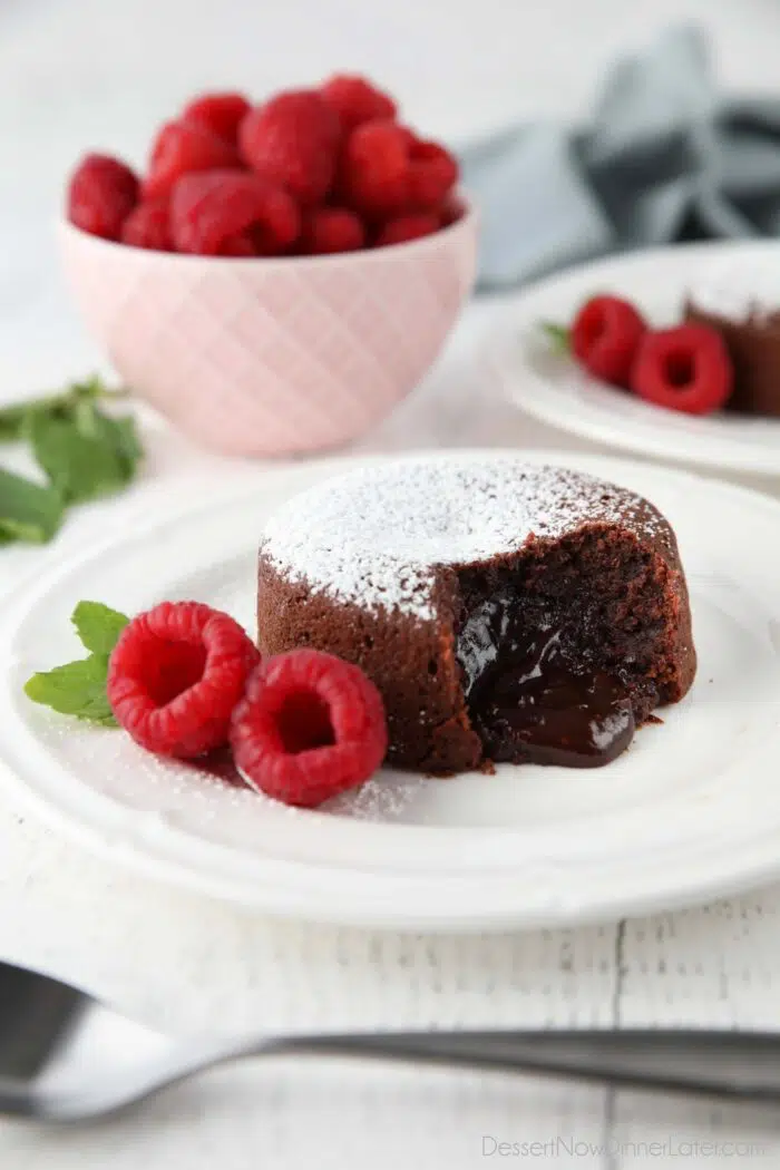 Molten chocolate lava cake on plate dusted with powdered sugar and a side of raspberries and mint, with gooey center melting onto the plate.
