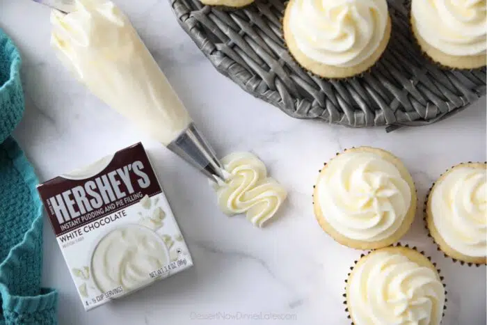 A box of Hershey's white chocolate instant pudding mix with frosting piped next to it and frosting piped on top of vanilla cupcakes.
